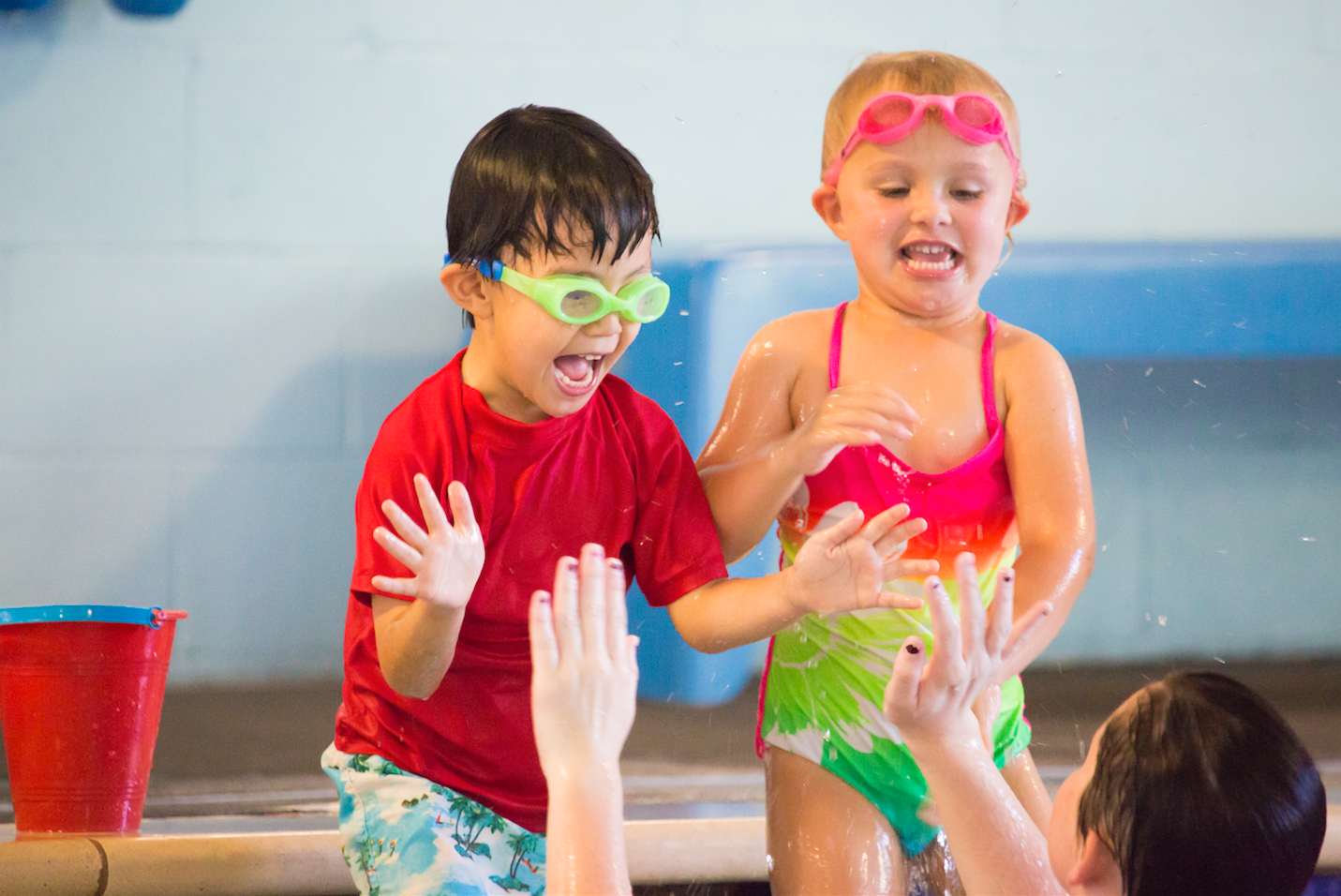 Two children in swimwear at a pool are splashing and laughing.  A woman in the water plays with them.