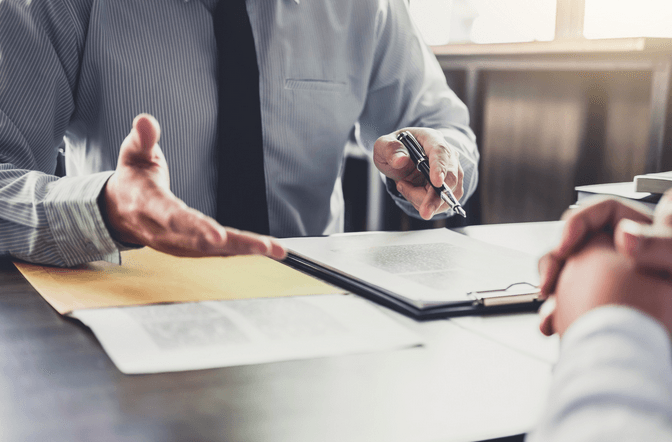 A man is sitting at a table talking to another man while holding a pen.