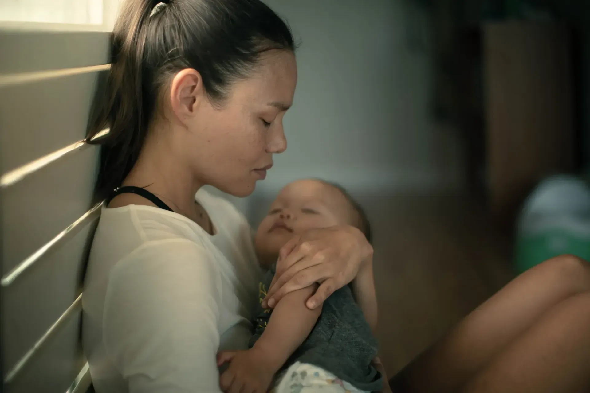 A woman is holding a baby in her arms while leaning against a wall.