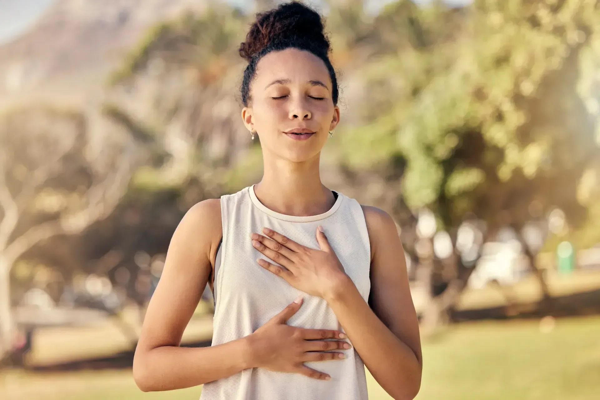 A woman is meditating in a park with her eyes closed and her hands on her chest.