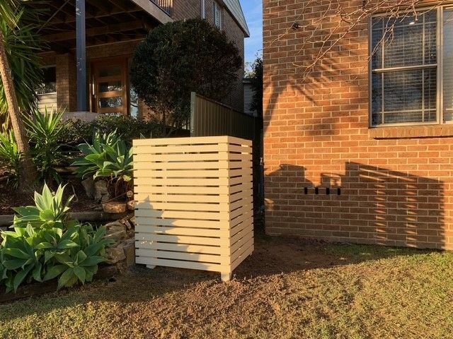 A wooden trash can is sitting in front of a brick house.