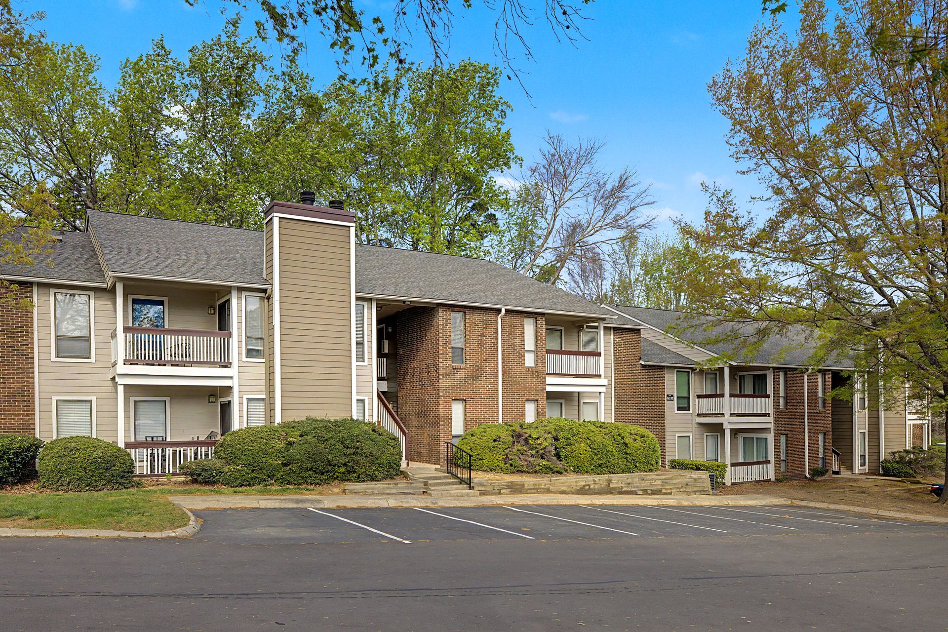 Apartment complex with tan siding, balconies, and trees along a parking lot