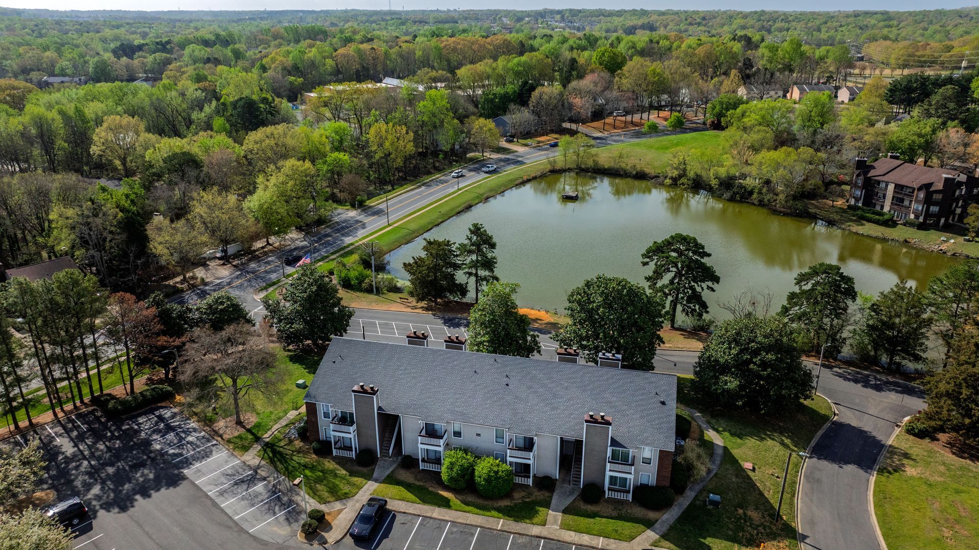 Aerial view of a pond beside a gray apartment building surrounded by trees and roads.