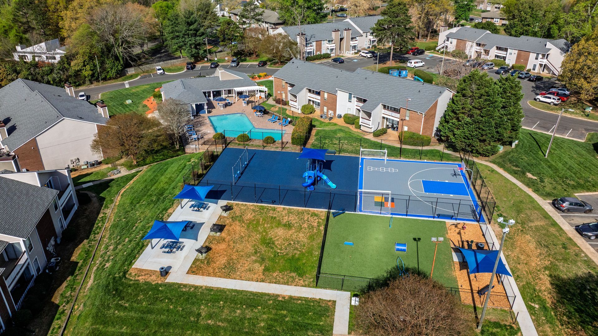 Aerial view of a backyard with a blue tennis court and swimming pool surrounded by houses and trees