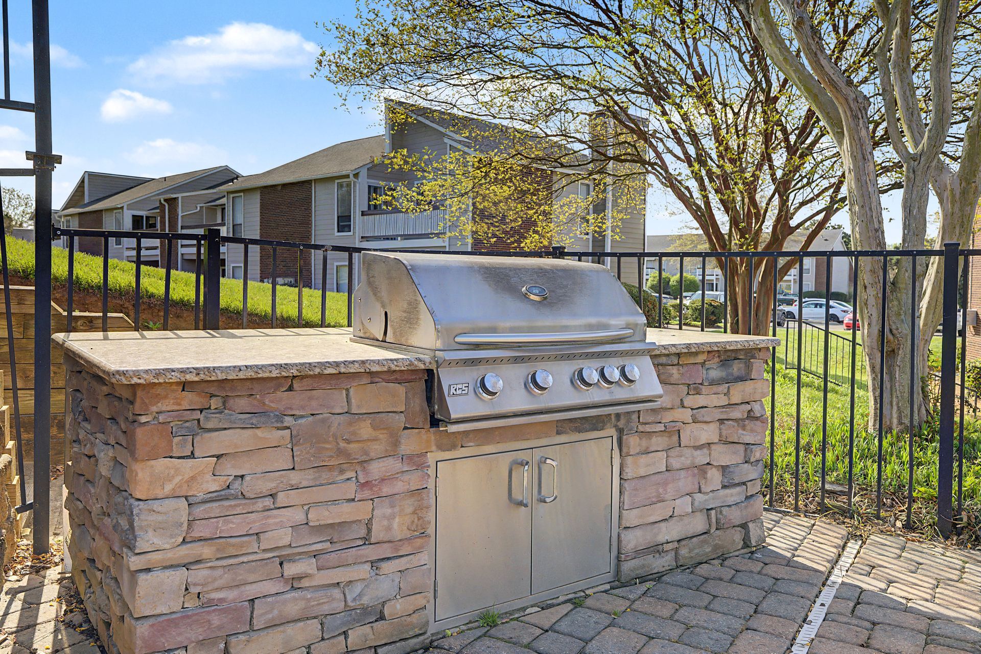 Built-in stainless steel grill beside a black fence in a sunny backyard patio.
