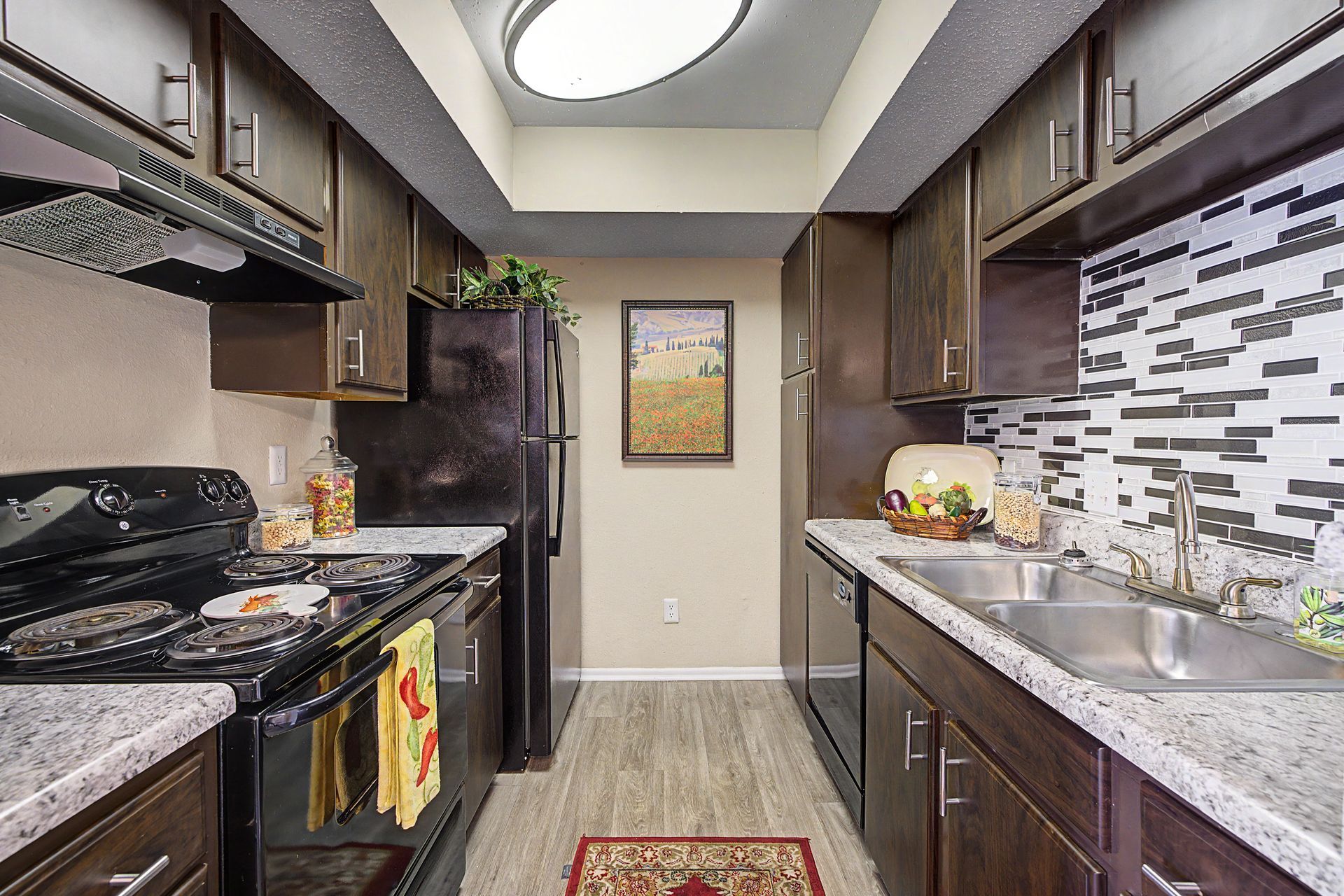 Narrow galley kitchen with dark cabinets, granite counters, mosaic backsplash, and black appliances