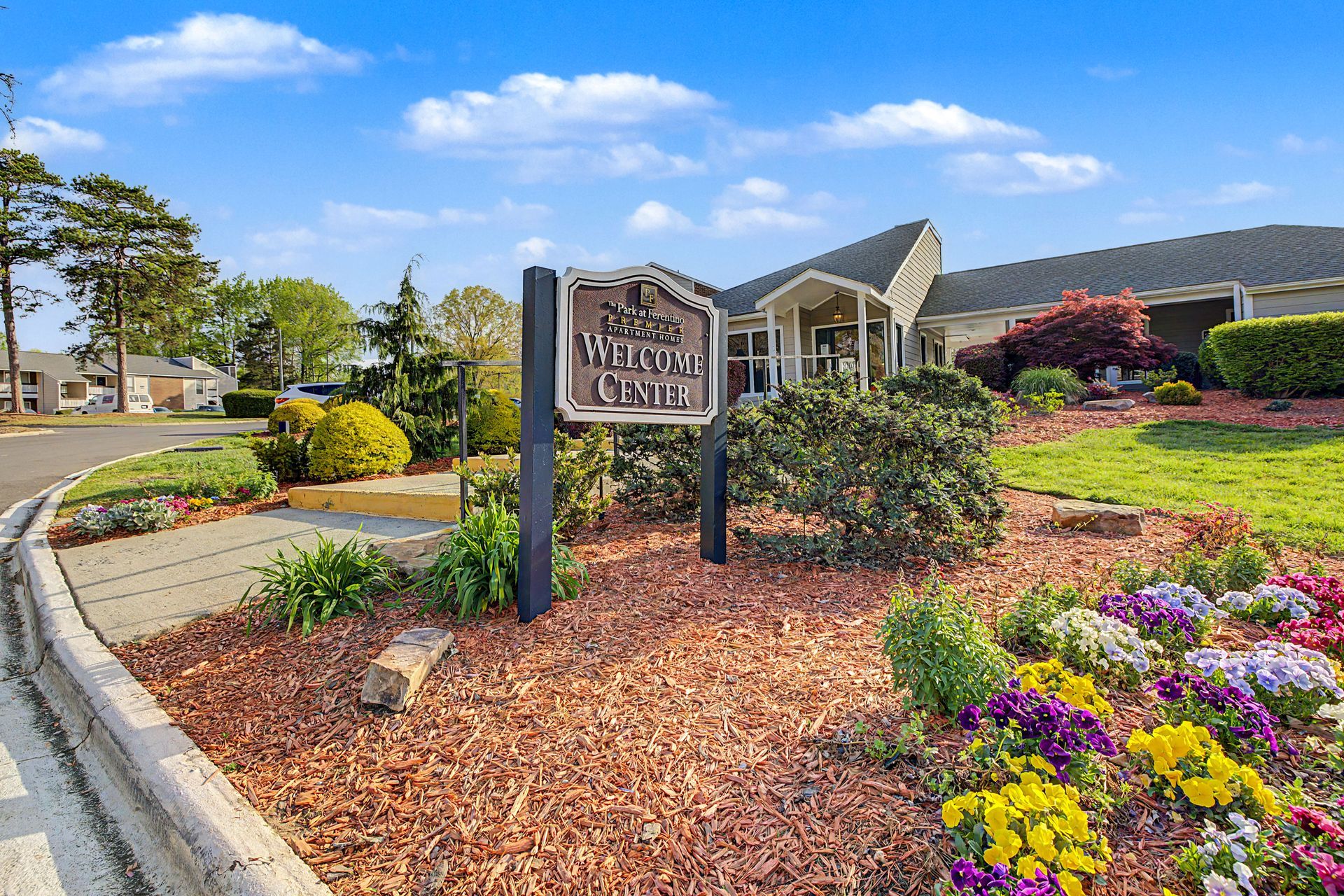 Landscaped apartment complex entrance with sign, flowers, shrubs, and brick buildings under a bright sky