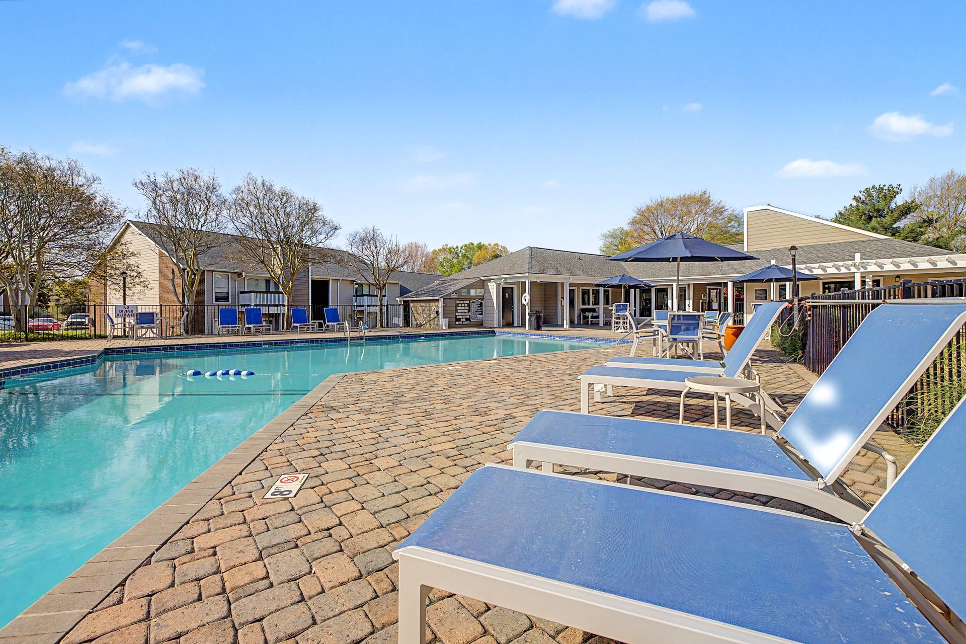 Resort pool with blue lounge chairs on a sunny patio beside low buildings and trees