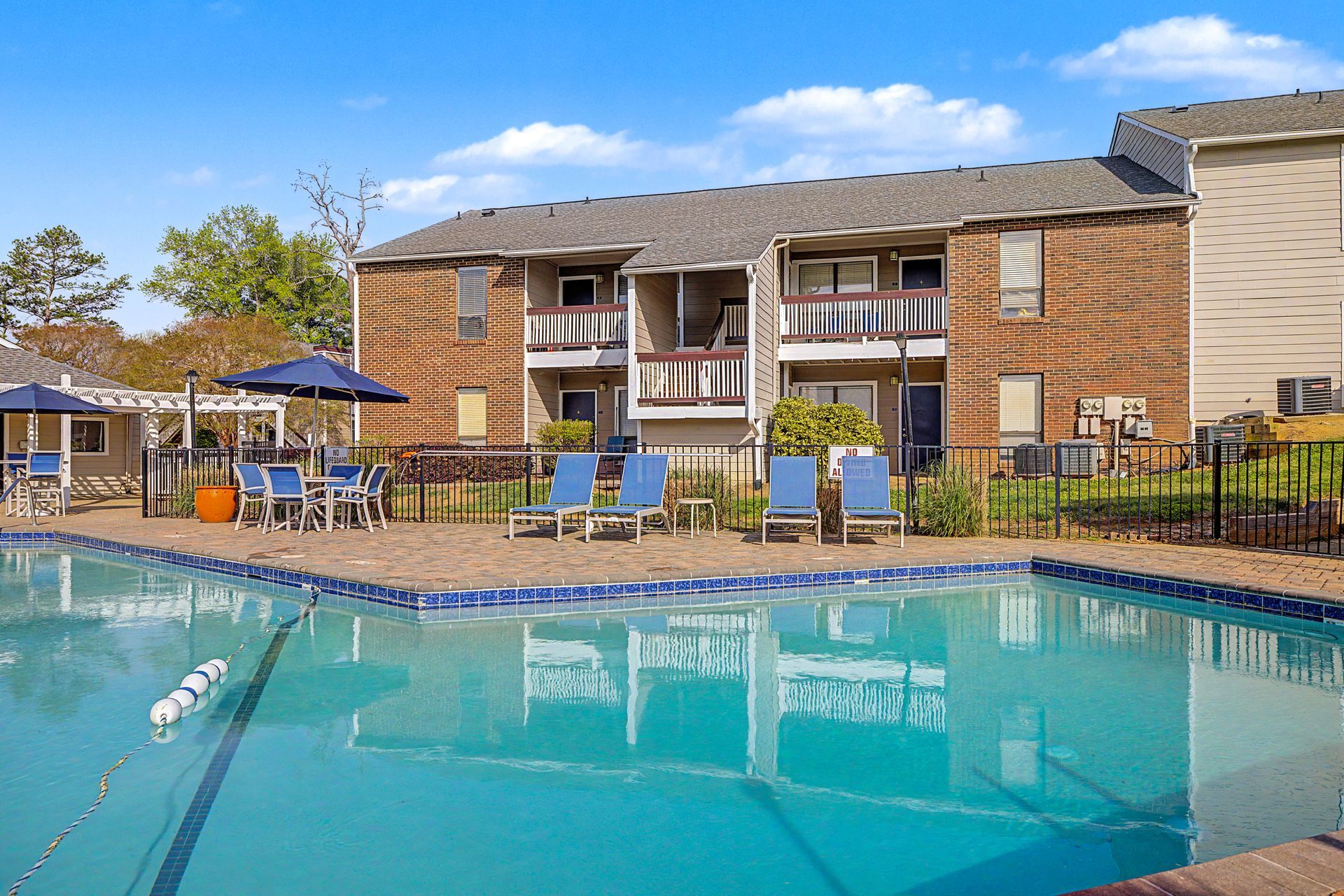 Apartment complex pool with blue water, lounge chairs, and patio umbrellas under a clear sky