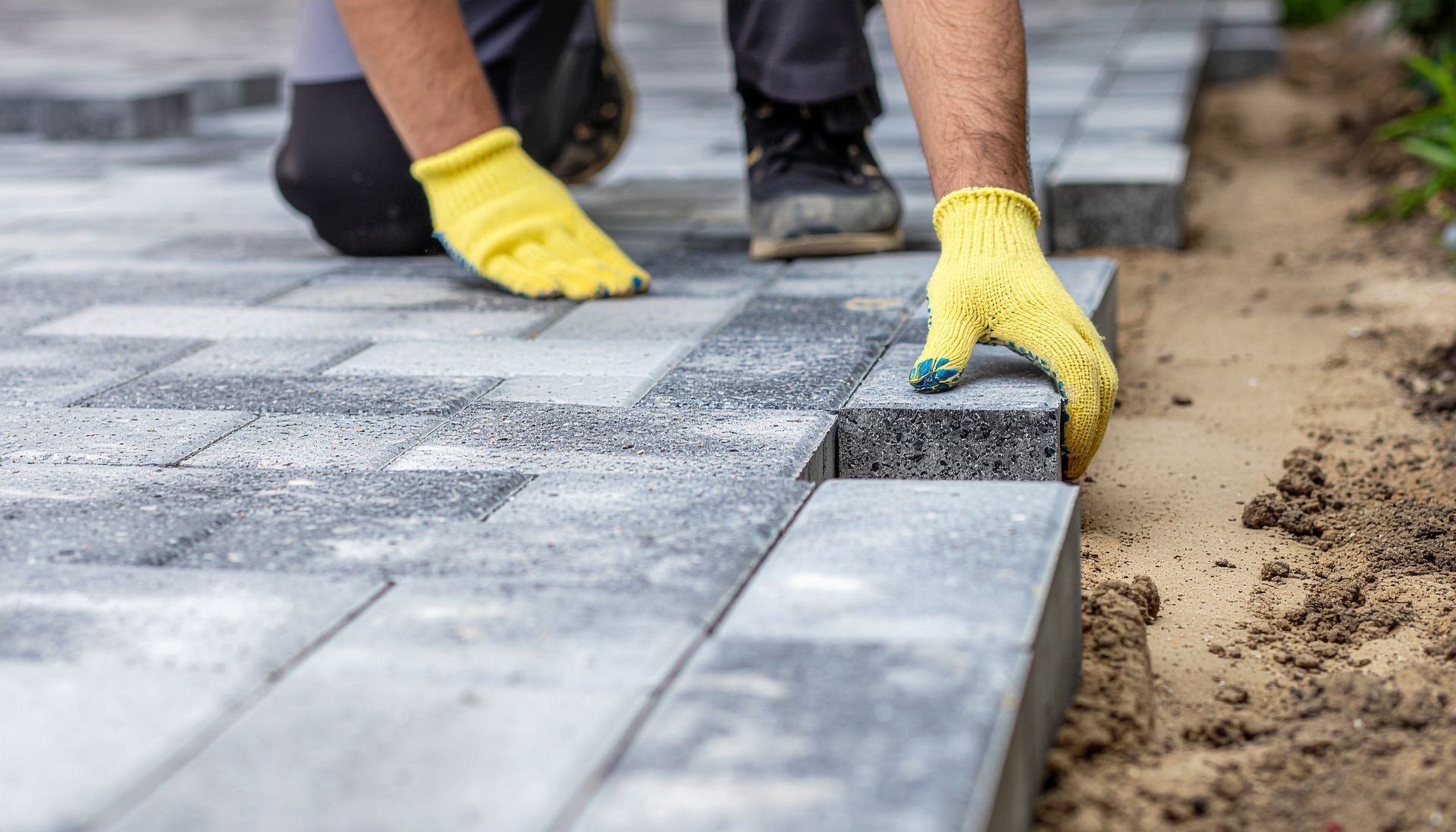 The master in work gloves lays paving stones in layers. Garden brick pathway paving by professional paver worker. Laying gray concrete paving slabs in house courtyard on sand foundation base. The master in work gloves lays paving stones in layers. Garden brick pathway paving by professional paver worker. Laying gray concrete paving slabs in house courtyard on sand foundation base.