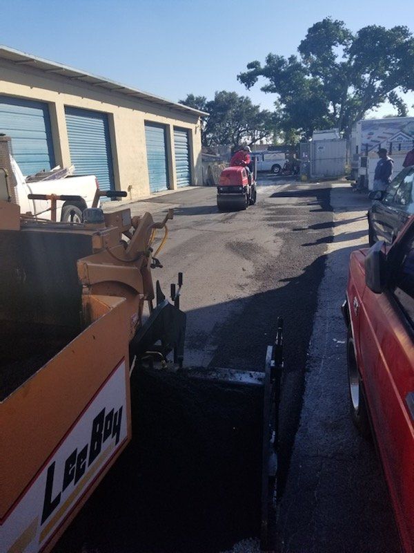 Asphalt paving in progress: a machine lays the blacktop, compacting roller behind it near a building.