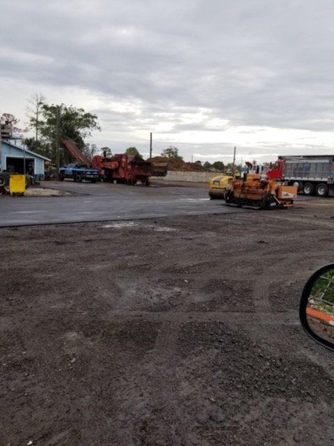 Construction site with asphalt paving equipment; trucks, machines, and building under overcast sky.