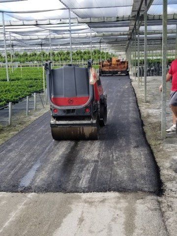 Asphalt paving machine compacting a freshly laid dark gray road inside a greenhouse; person stands to the side.