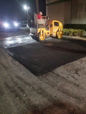 A person operating a yellow road roller compacts fresh asphalt in a dark parking lot at night.