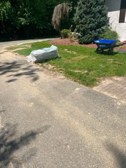 Asphalt driveway with a bag, blue wheelbarrow, and grass with a tree in the background.