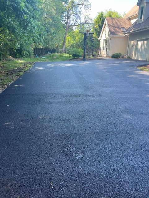 Newly paved asphalt driveway leading to a beige house, basketball hoop in the background, surrounded by trees.