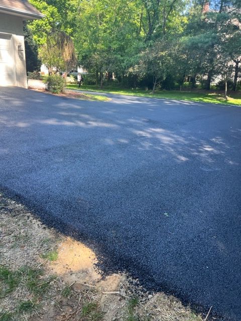 Freshly paved asphalt driveway in front of a house, leading to a grassy yard with trees.