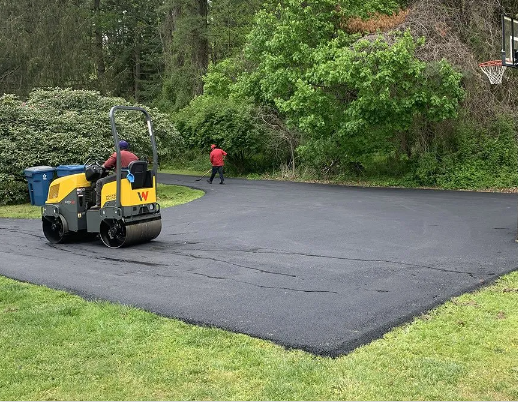 Workers paving a dark asphalt driveway beside a yellow roller in a green wooded area. Workers paving a dark asphalt driveway beside a yellow roller in a green wooded area.
