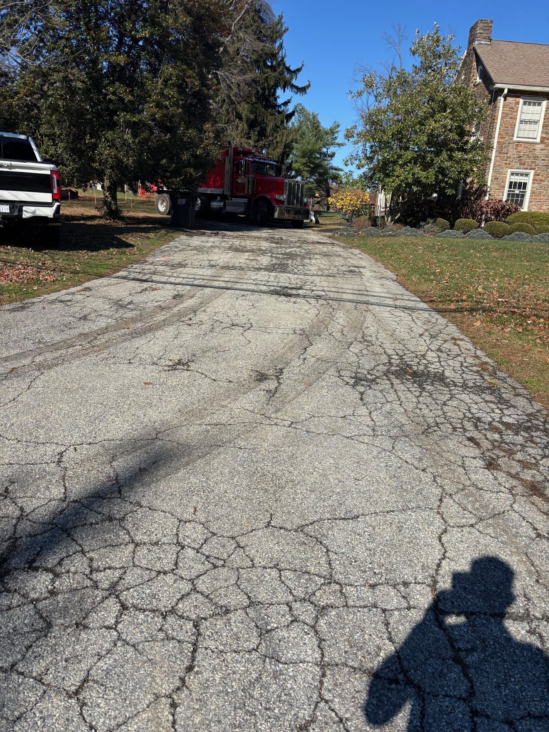 Cracked driveway leading to a house, with parked vehicles and trees in autumn sunlight.