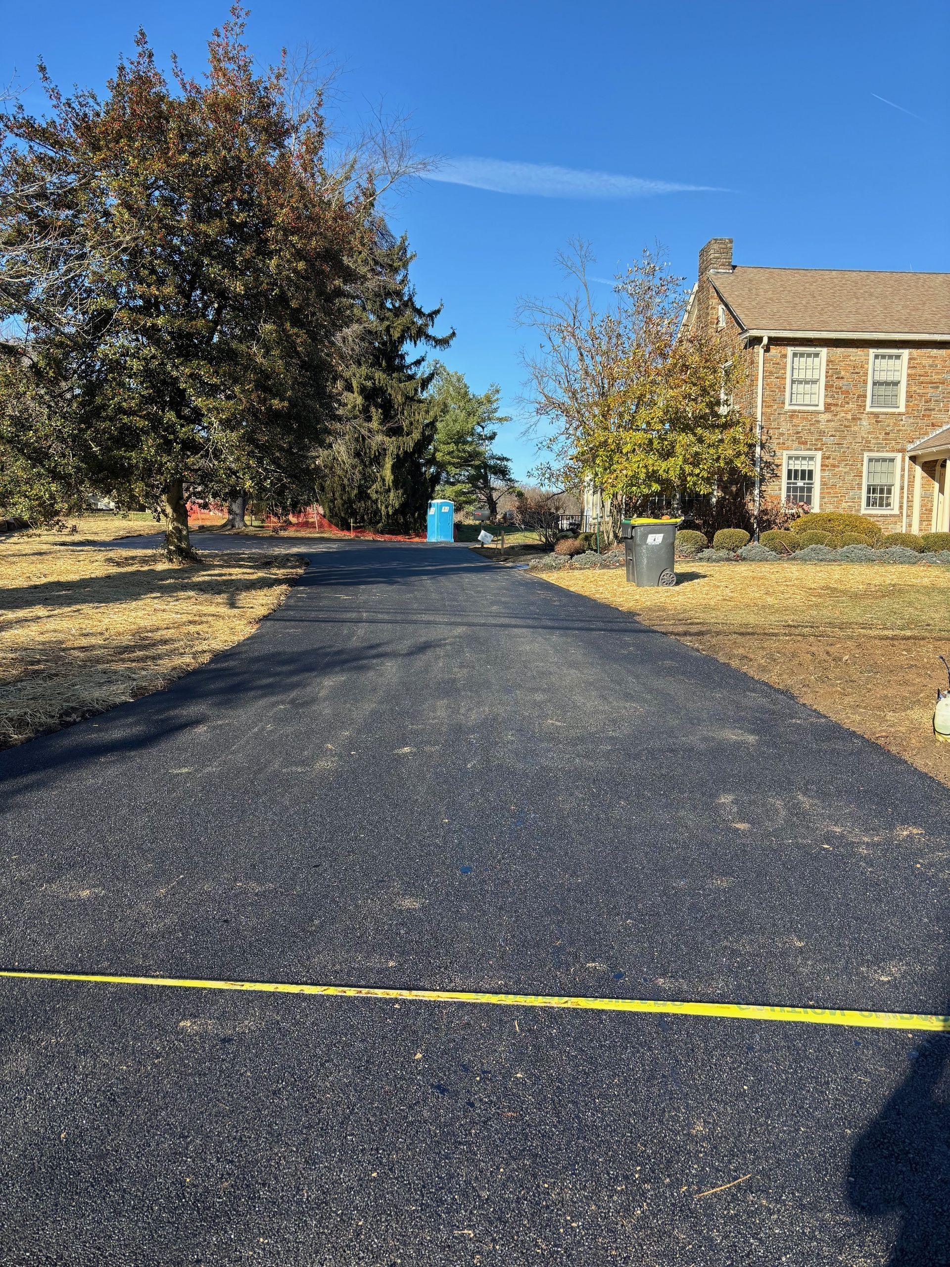 New blacktop driveway leading to a stone house, with autumn trees and blue sky.
