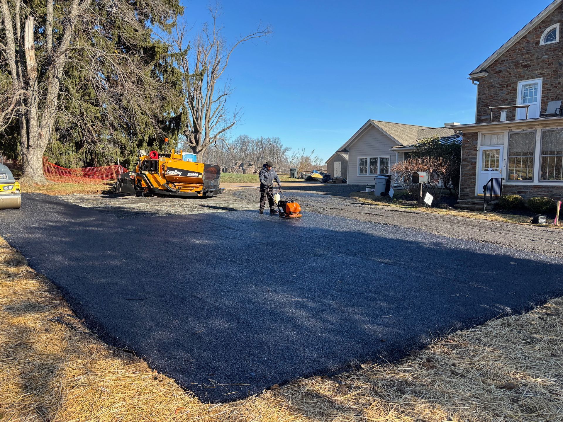 Crew paving a driveway beside a house, with a roller and worker on a sunny day