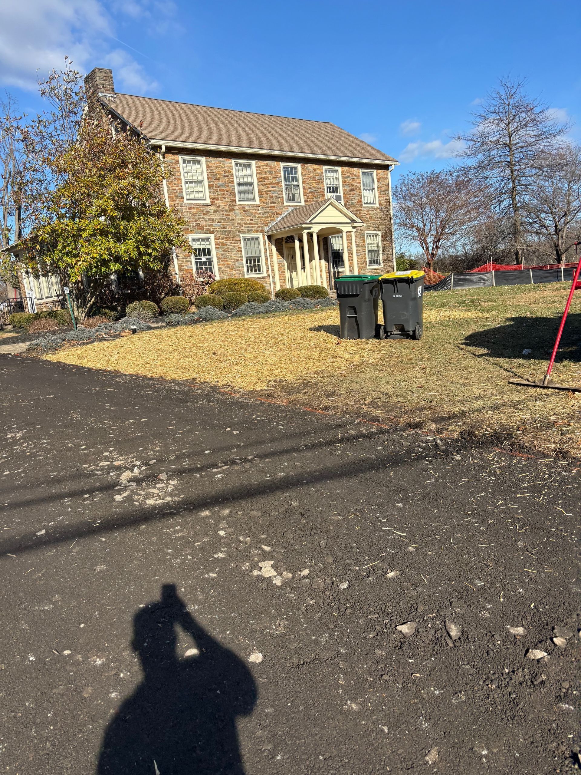 Stone house with a tiled roof and two trash bins in a leaf-covered yard on a sunny day