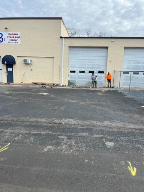 Building with garage doors and two workers standing outside. Paved area with arrows. Cloudy sky.
