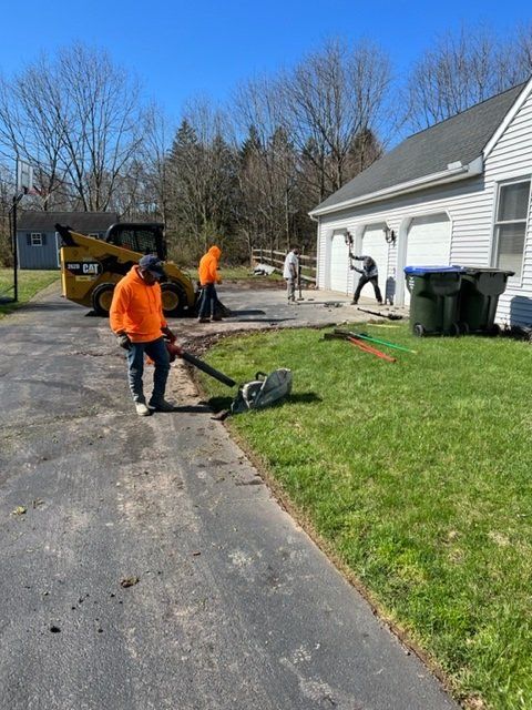 Three workers in orange vests use tools near a house and driveway. A yellow skid steer loader is also present.