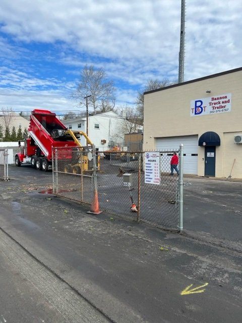 Red dump truck unloading asphalt near a building, next to a fence. A person in a red jacket walks by.