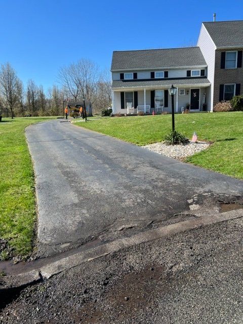 Asphalt driveway in front of a two-story house with grass, a worker, and a clear sky.