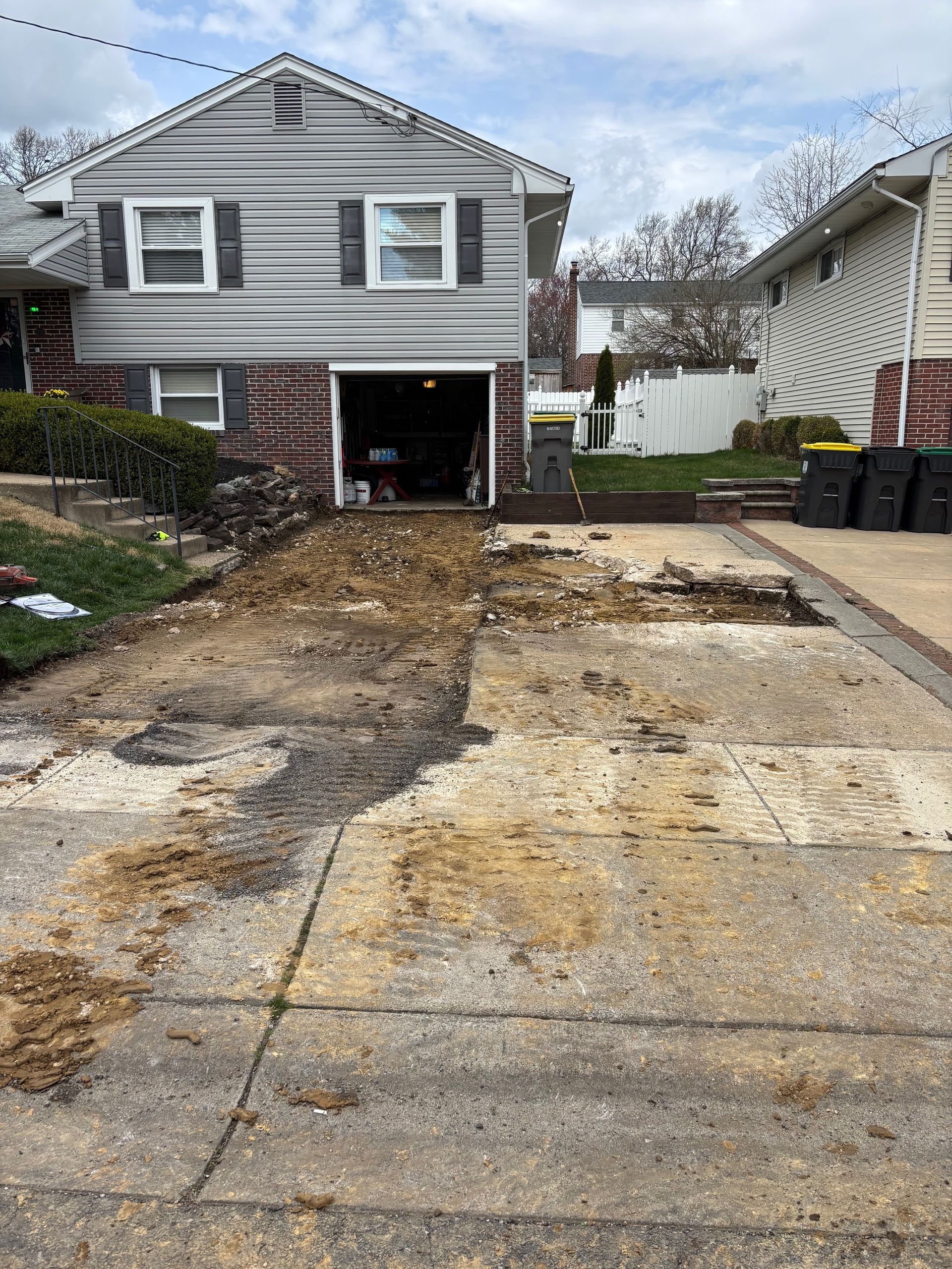 Driveway demolition in front of a two-story house with a garage. Dirt and debris cover the driveway.
