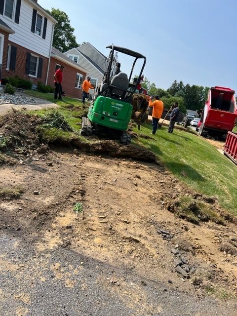 Mini excavator and crew digging on a residential lawn, next to a driveway and street, on a sunny day.