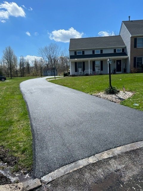 Newly paved black asphalt driveway curves towards a two-story house with green lawn on a sunny day.