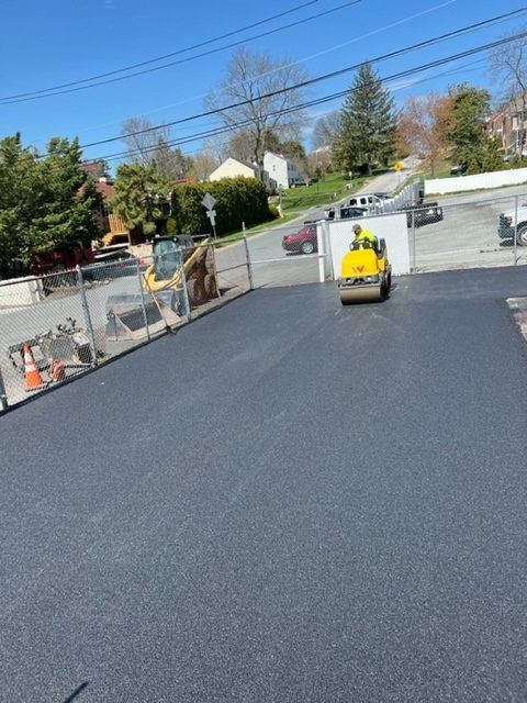 Asphalt parking lot being rolled by a yellow compactor machine. Fenced in with construction equipment nearby. Sunny day.