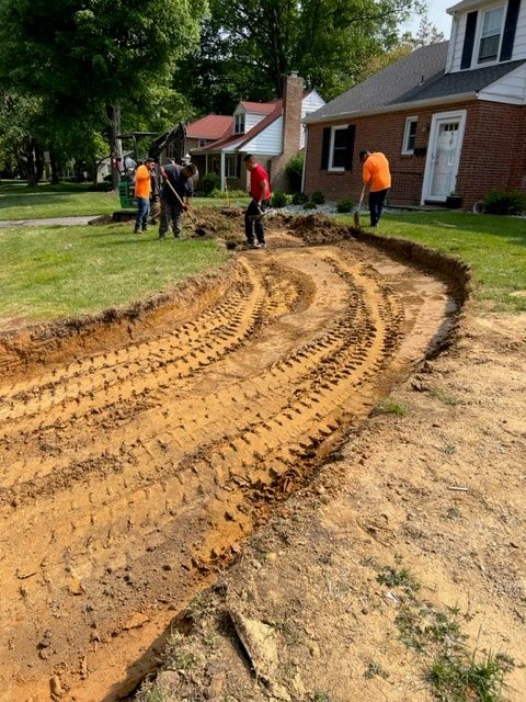 Construction workers dig a trench along a residential lawn, marked by tractor tire tracks, houses in background.