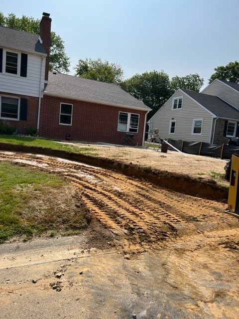 Construction site with trench in front of brick and siding houses, dirt, and tire tracks.