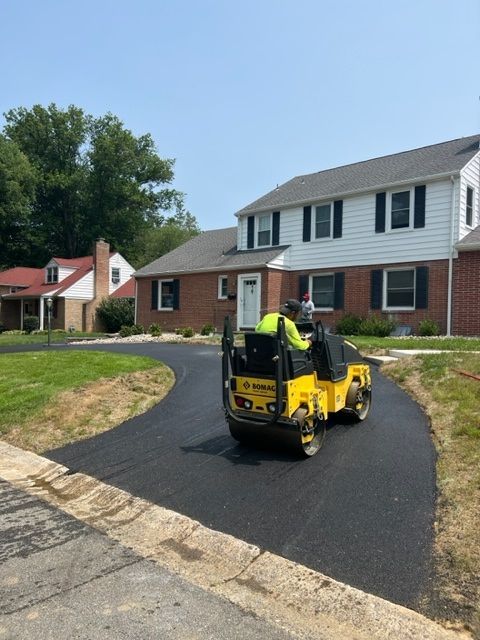 A yellow asphalt roller compacting fresh asphalt driveway in front of a brick house.
