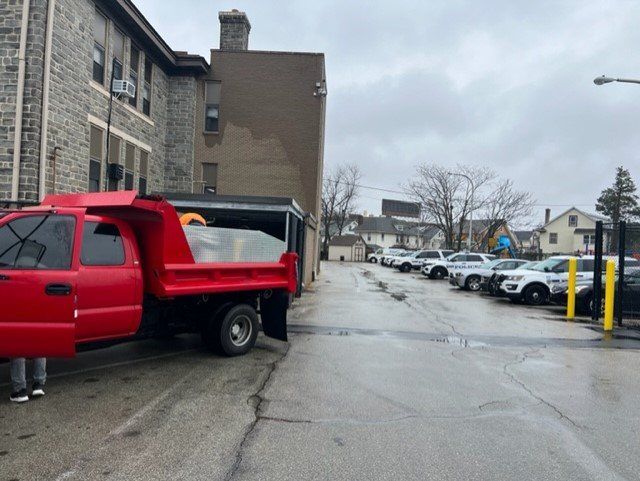 Red dump truck parked near a building with multiple police cars in the background. Overcast day.