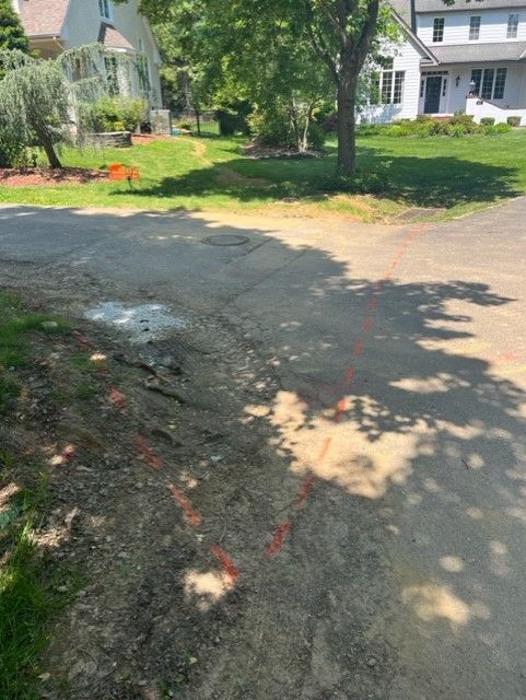 Dirt road with orange paint markings, green grass, and houses in the background on a sunny day.