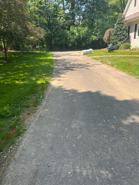 Asphalt driveway with grass on either side, leading toward trees and a house. Sunny day.