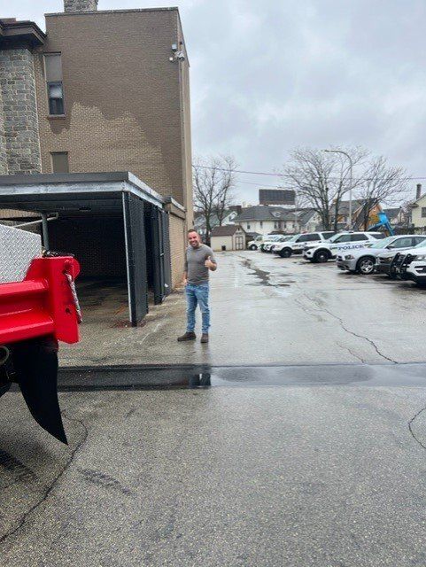 Man giving a thumbs up next to a building and cars on a cloudy day.