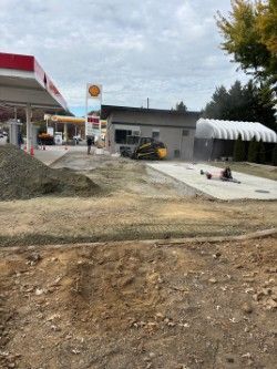 Gas station construction site with Shell branding. Excavator and workers present. Cloudy sky.