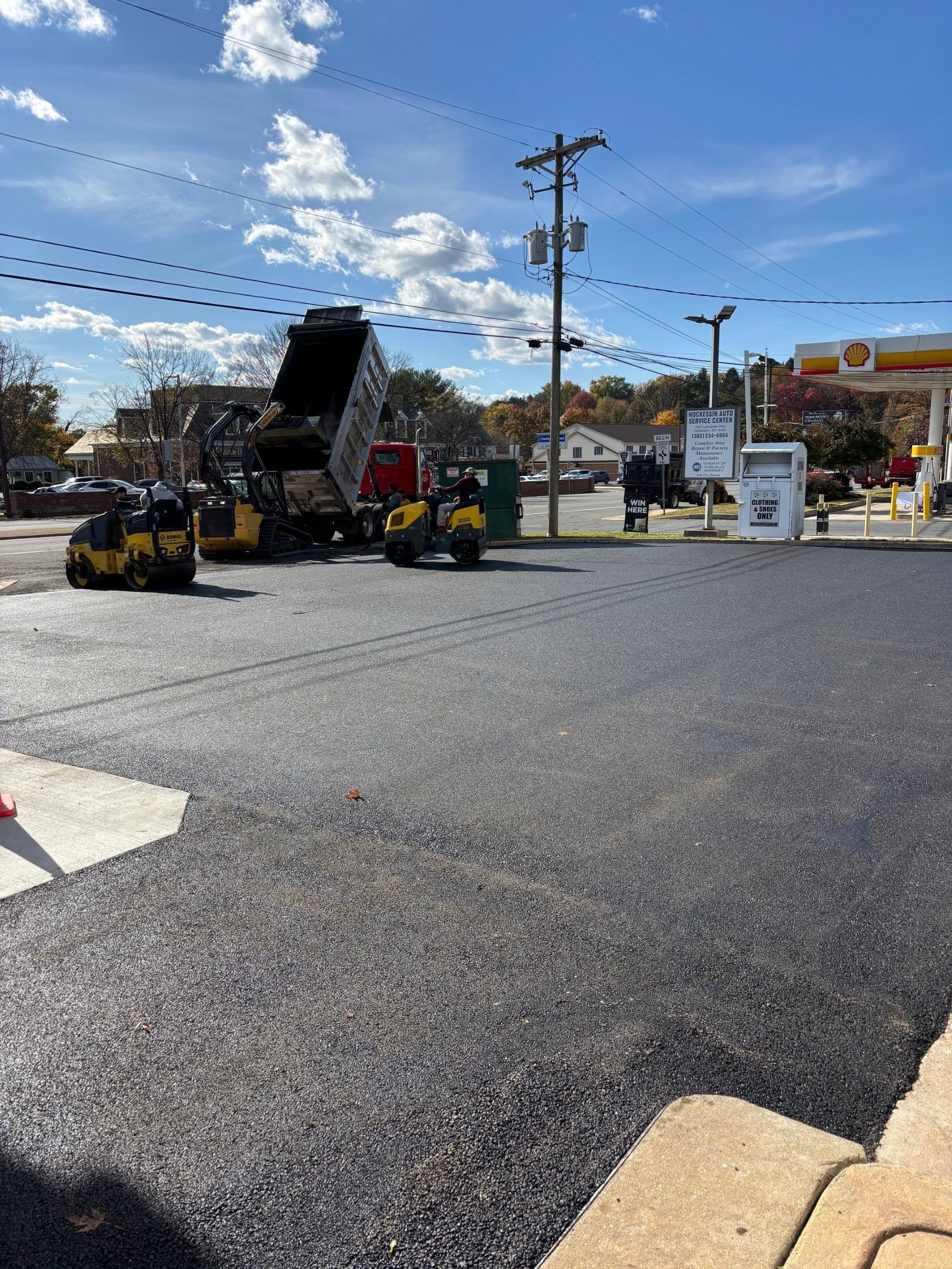 Asphalt paving in progress at gas station; dump truck, rollers, workers, clear sky.