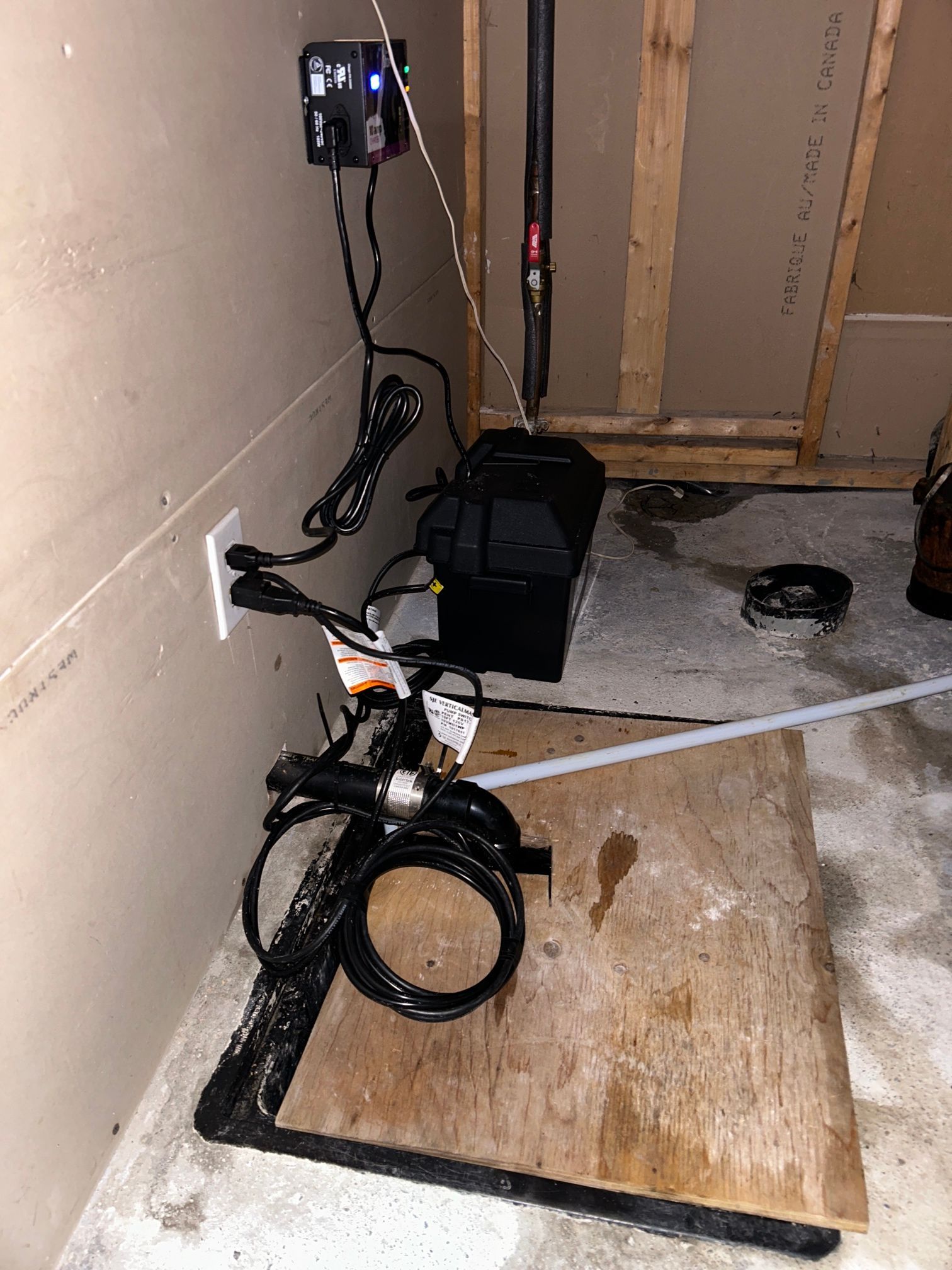 Un aménagement de sous-sol comprenant une pompe de puisard et un système de batterie de secours sur une plate-forme en bois, avec des cordons électriques et des tuyaux visibles.