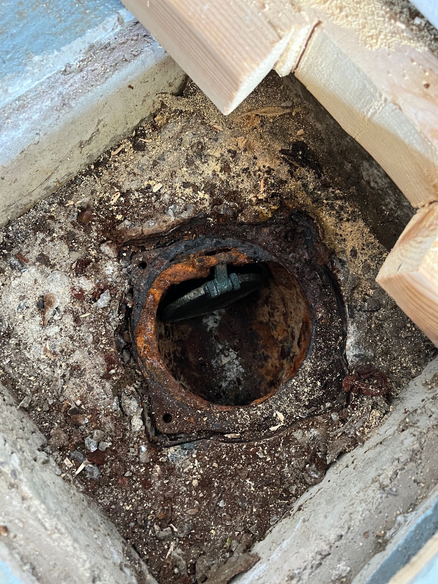 Tuyau d'évacuation corrodé dans un socle en béton, entouré de bois et de débris. Le métal est rouillé.