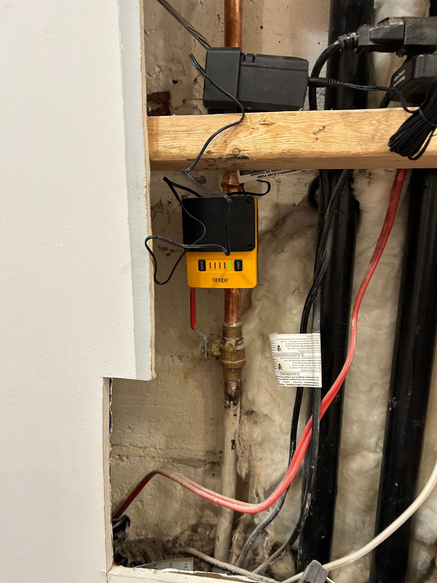 Vanne d'eau intelligente jaune installée sur un tuyau en cuivre, montée sur une poutre en bois, avec divers fils et tuyaux visibles dans une cavité murale.