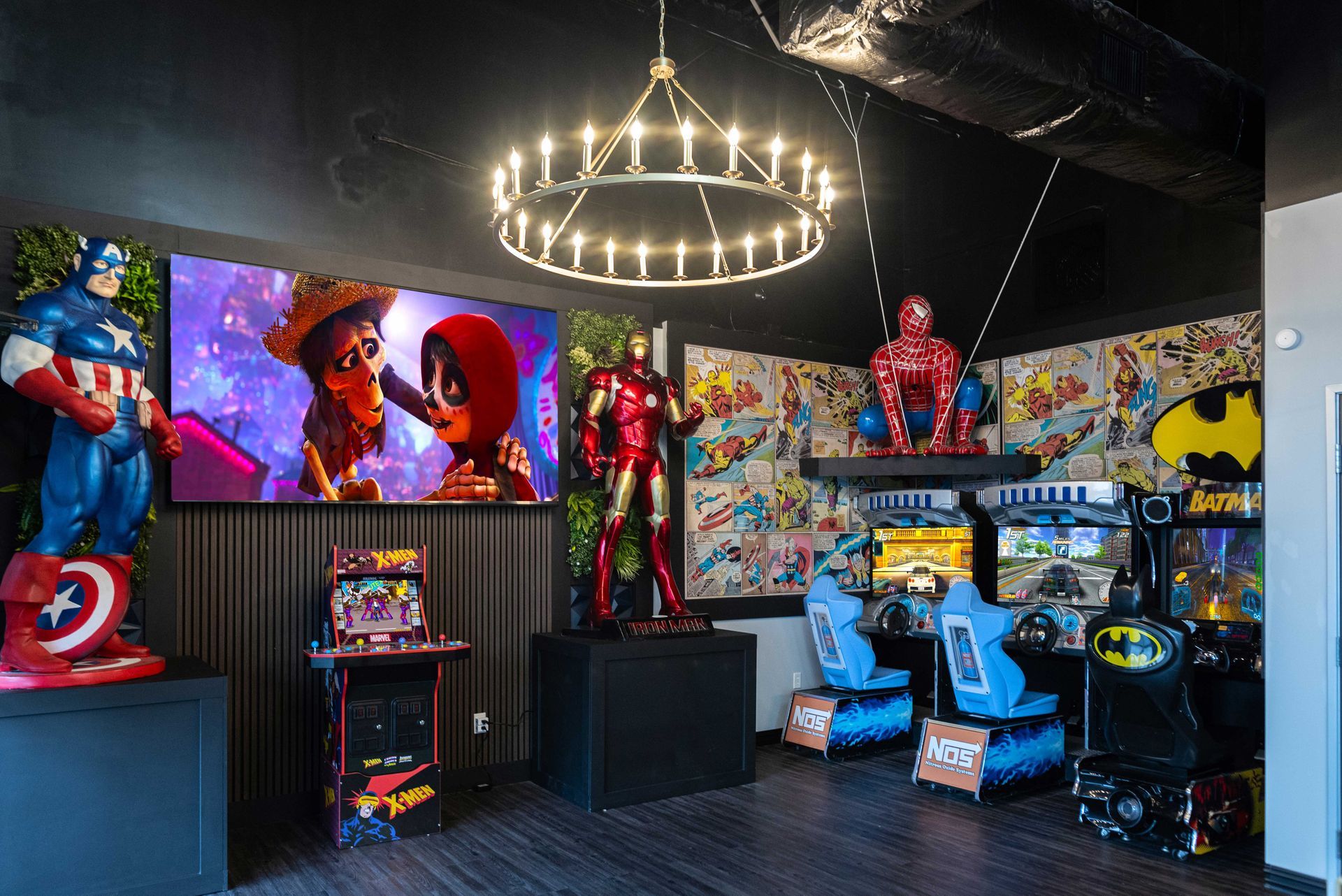 Marvel-themed arcade room with superhero statues, pinball machines, and wall art under chandelier lighting