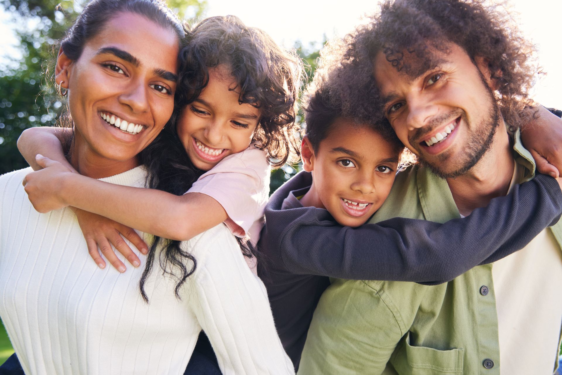 Two smiling adults with two children hugging outdoors in bright sunlight