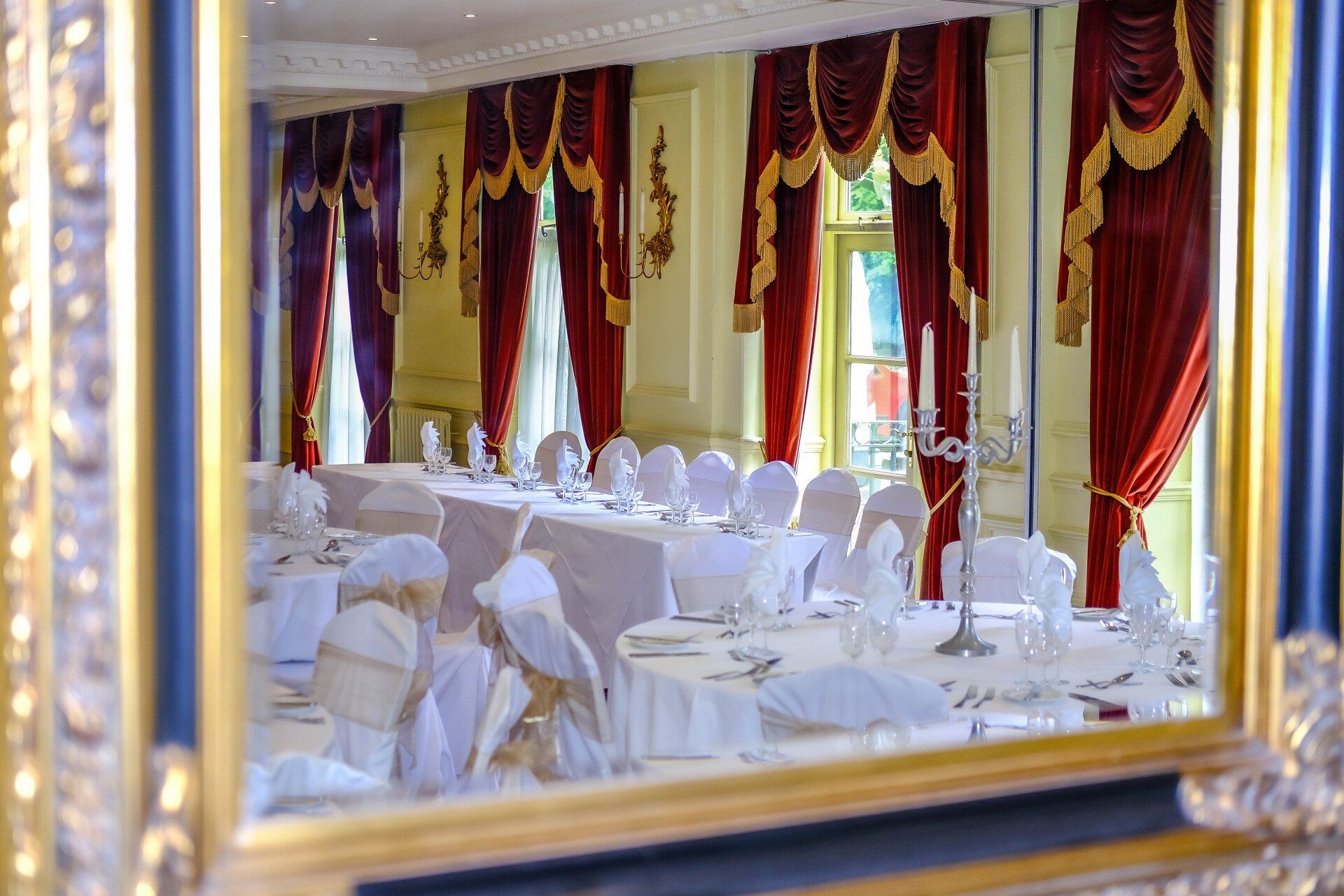 A room with tables and chairs set up for a wedding reception is reflected in a mirror.
