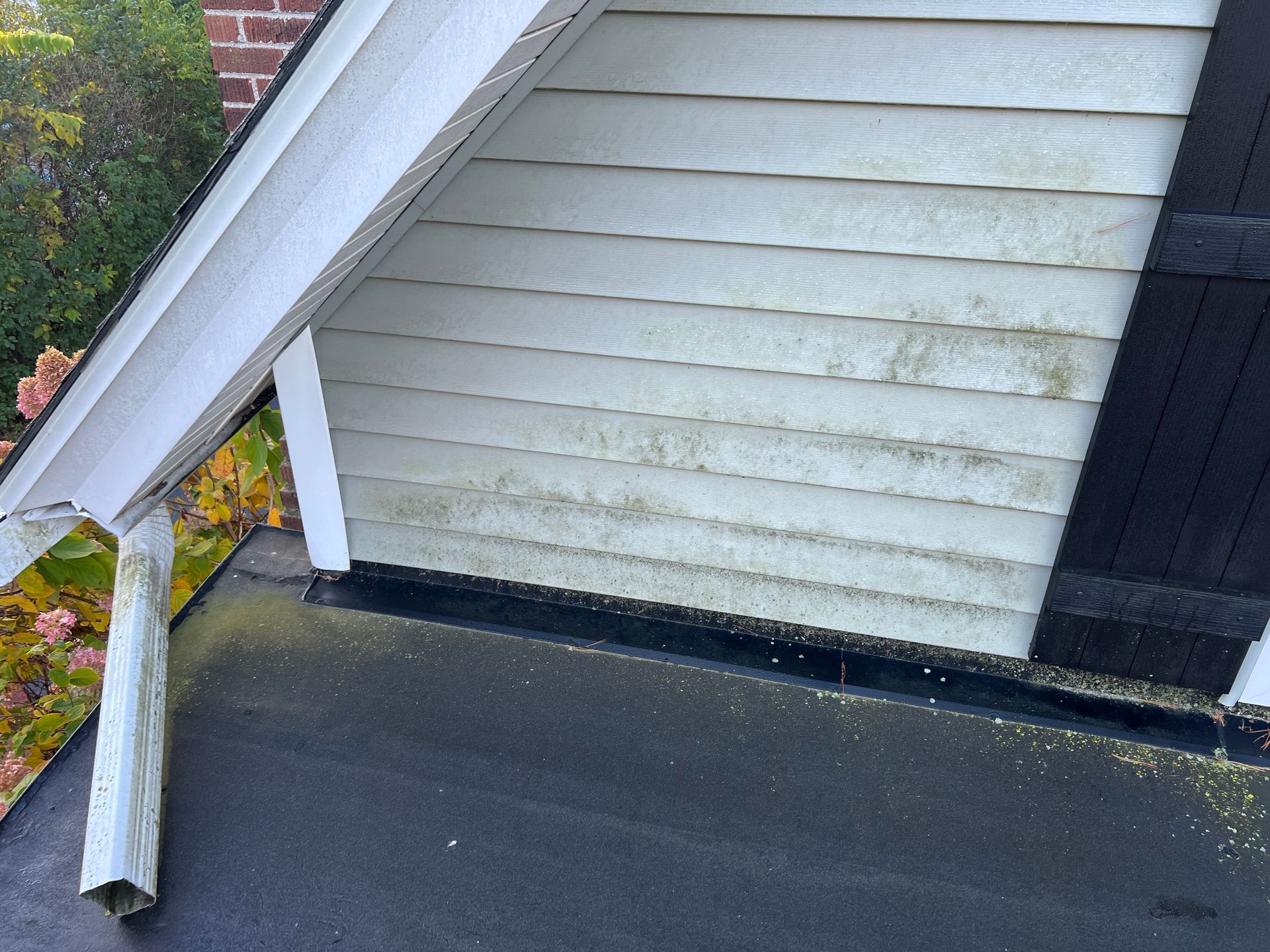 White siding with grime buildup above a black roof section, framed by a roof eave and dark shutter.